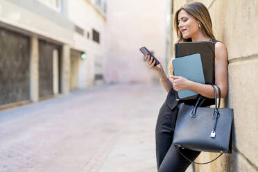 Geschäftsfrau mit Handtasche, die ein Smartphone benutzt und sich an die Wand lehnt - DLTSF03690