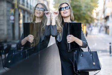 Lächelnde Geschäftsfrau mit Sonnenbrille, die sich an eine Glaswand lehnt - DLTSF03695