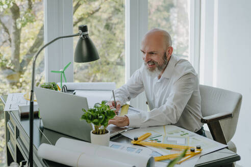 Glücklicher reifer Architekt arbeitet am Laptop mit Blaupausen am Schreibtisch - YTF01429
