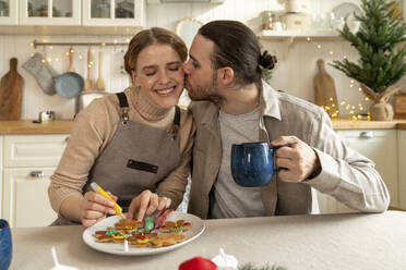 Mann küsst Frau beim Verzieren von Keksen am Tisch - VPIF08935