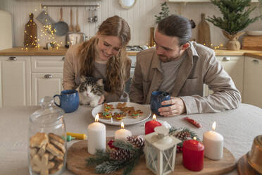 Smiling woman holding cat with man sitting at table - VPIF08948