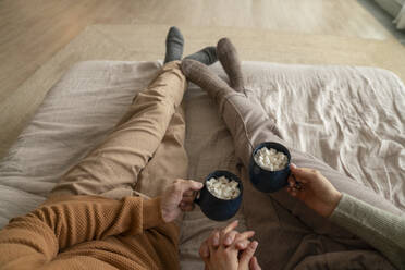 Couple holding mugs of hot chocolate with marshmallows on bed at home - VPIF08962