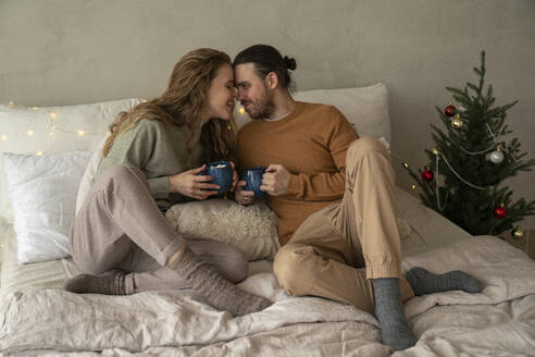 Romantisches Paar hält Tassen auf dem Bett in der Nähe von Weihnachtsbaum zu Hause - VPIF08963
