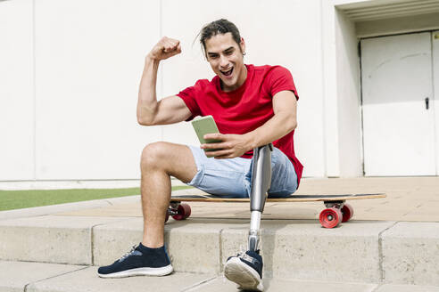 Fröhlicher Mann mit Behinderung, der seine Muskeln anspannt und ein Mobiltelefon benutzt - JCZF01299