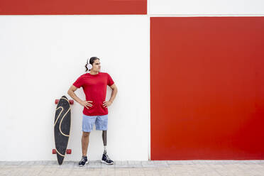 Junger Mann mit drahtlosen Kopfhörern und Beinprothese in der Nähe eines Skateboards vor einer Wand stehend - JCZF01301