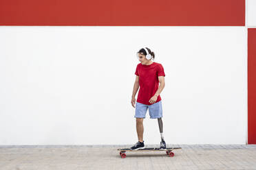 Junger Mann mit drahtlosen Kopfhörern und Skateboarding mit Beinprothese vor einer Wand - JCZF01305