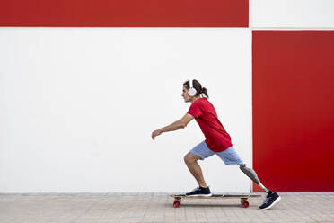 Mann mit drahtlosen Kopfhörern und Skateboarding mit Beinprothese vor einer Wand - JCZF01306