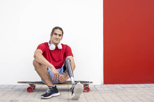 Smiling young man sitting on skateboard with prosthetic leg in front of wall - JCZF01309