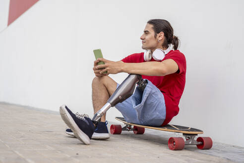 Lächelnder Mann mit Behinderung sitzt auf einem Skateboard und hält ein Smartphone - JCZF01312