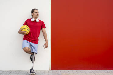 Lächelnder Mann mit Behinderung, der einen gelben Basketball hält und sich an die Wand lehnt - JCZF01314