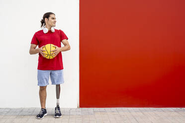 Lächelnder junger Mann mit Beinprothese, der einen gelben Basketball vor einer Wand hält - JCZF01316