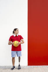 Glücklicher junger Mann mit Beinprothese hält gelben Basketball vor einer Wand - JCZF01317