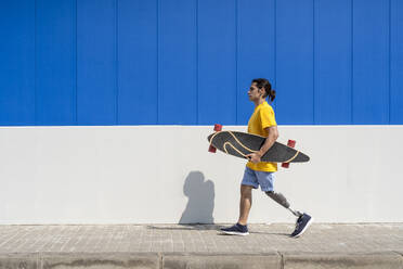 Junger Mann hält Skateboard und geht mit Beinprothese vor einer Wand - JCZF01320