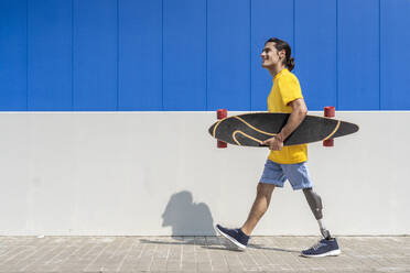 Glücklicher junger Mann, der ein Skateboard hält und mit einer Beinprothese vor einer Wand läuft - JCZF01321