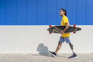 Happy young man holding skateboard and walking with prosthetic leg in front of wall - JCZF01321