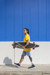 Smiling young man holding skateboard and walking with prosthetic leg in front of wall - JCZF01322