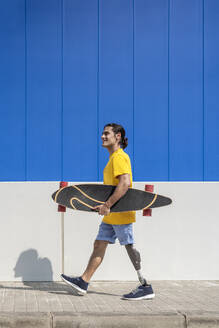 Lächelnder junger Mann, der ein Skateboard hält und mit einer Beinprothese vor einer Wand läuft - JCZF01322