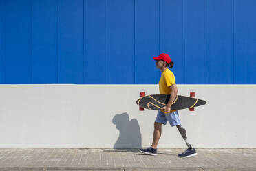 Man holding skateboard and walking with prosthetic leg in front of wall - JCZF01323