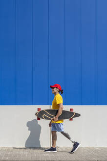 Man with disability holding skateboard and walking near wall - JCZF01324