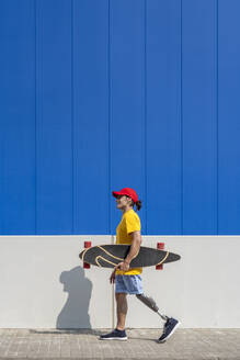 Mann mit Behinderung hält Skateboard und geht in der Nähe der Wand - JCZF01324