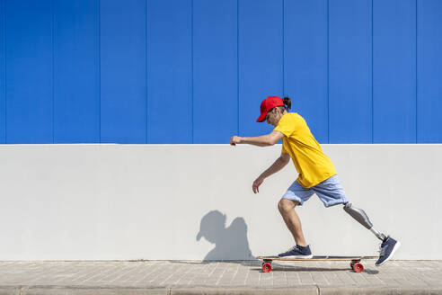Junger Mann mit Behinderung fährt Skateboard in der Nähe einer Mauer - JCZF01325