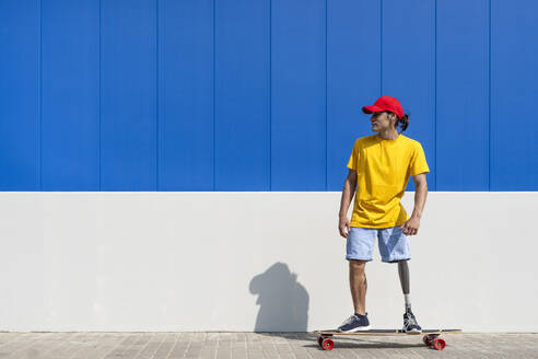 Junger Mann mit Behinderung steht auf einem Skateboard in der Nähe einer Mauer - JCZF01326