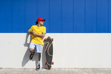 Junger Mann mit Behinderung hält Smartphone und steht neben Skateboard an einem sonnigen Tag - JCZF01330