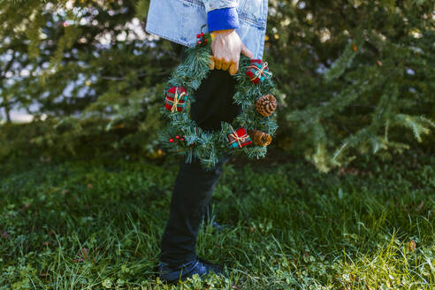 Man holding Christmas wreath and standing on grass near tree - EGHF00802
