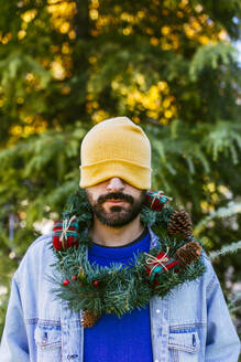 Mann bedeckt Augen mit Strickmütze und trägt Weihnachtskranz um den Hals - EGHF00803