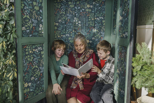 Urgroßmutter liest ihren Enkeln zu Hause im Schrank sitzend ein Buch vor - VBUF00517