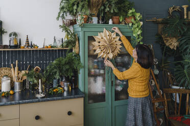 Ältere Frau hängt Papierschneeflocke auf Vitrine in Küche zu Hause - VBUF00519