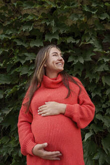Lächelnde schwangere Frau im roten Kleid und vor Pflanzen stehend - AAZF01351