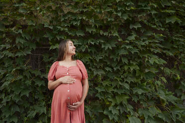Lächelnde schwangere Frau im Kleid und vor Pflanzen stehend - AAZF01353