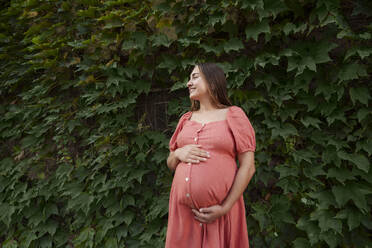 Glückliche schwangere Frau im Kleid und vor Pflanzen stehend - AAZF01354