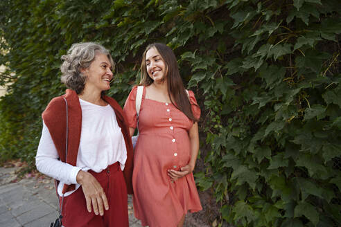 Lächelnde schwangere Tochter im Gespräch und beim Spaziergang mit der Mutter vor Pflanzen - AAZF01357