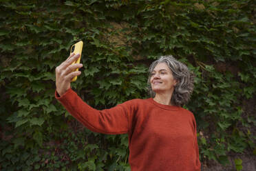 Smiling woman talking selfie with smart phone in front of plants - AAZF01374