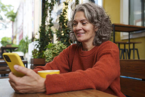 Glückliche reife Frau mit Smartphone und Kaffeetasse auf dem Tisch eines Straßencafés - AAZF01382