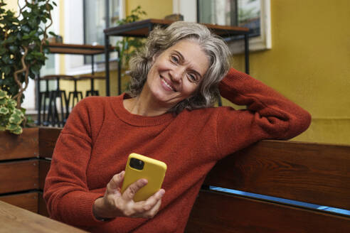 Smiling woman holding smart phone and sitting at sidewalk cafe - AAZF01386