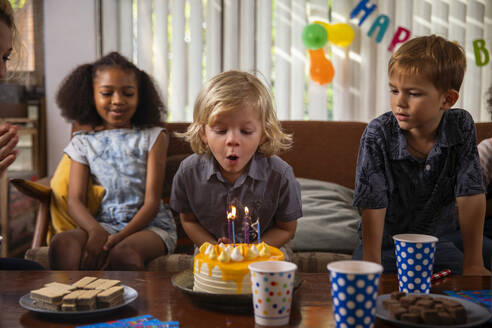 Junge bläst Kerzen auf seinem Geburtstagskuchen aus, während seine Freunde zusehen - IKF01432