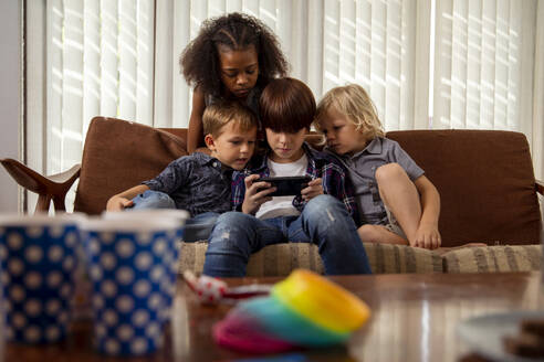Junge spielt Spiel auf Smartphone mit Freunden auf Couch sitzend - IKF01442
