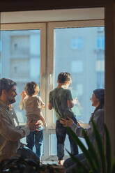 Parents holding children looking outside through window at home - ANAF02466