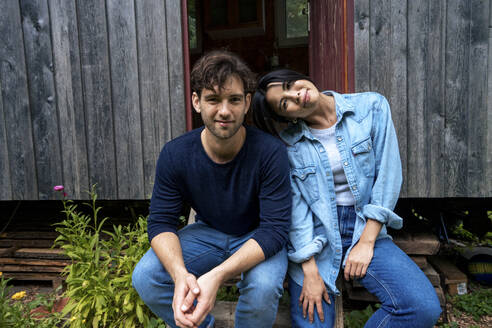 Happy woman leaning head on man sitting in front of log cabin - JOSEF22160