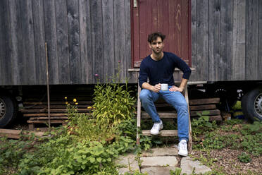 Lächelnder Mann sitzt mit einer Kaffeetasse auf den Stufen vor einer Blockhütte - JOSEF22175