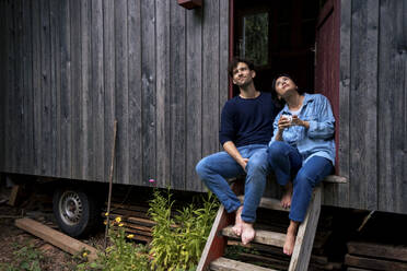 Smiling man and woman sitting on steps in doorway of log cabin - JOSEF22179