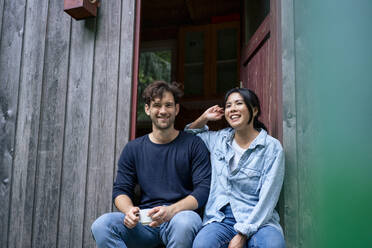 Happy man and woman sitting in doorway of log cabin - JOSEF22181
