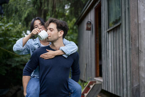 Woman with man drinking coffee through cup near log cabin - JOSEF22184