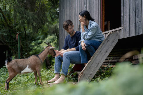 Woman sitting with man petting goat near wooden cabin - JOSEF22206