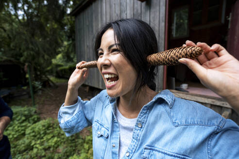 Cheerful woman holding pine cones over ears in front of wooden cabin - JOSEF22215