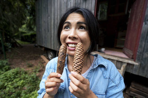 Happy woman holding pine cones in front of cabin - JOSEF22216