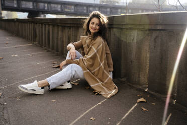 Lächelnde junge Frau sitzt auf dem Boden in der Nähe der Wand - YBF00318